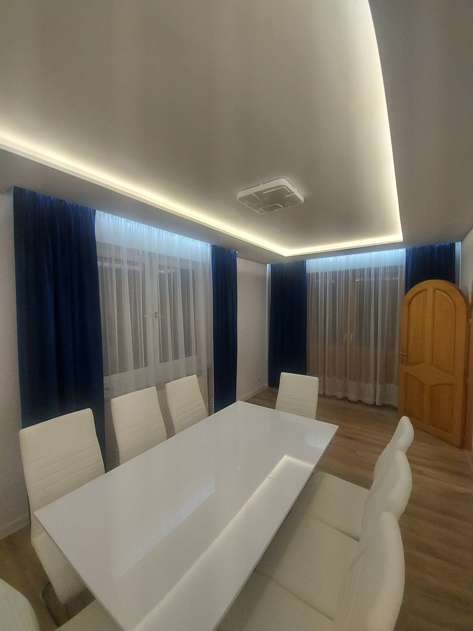 Nowoczesna jadalnia z białym stołem, białymi krzesłami i podświetlanym sufitem, okna z granatowymi i białymi zasłonami, drewniana podłoga.