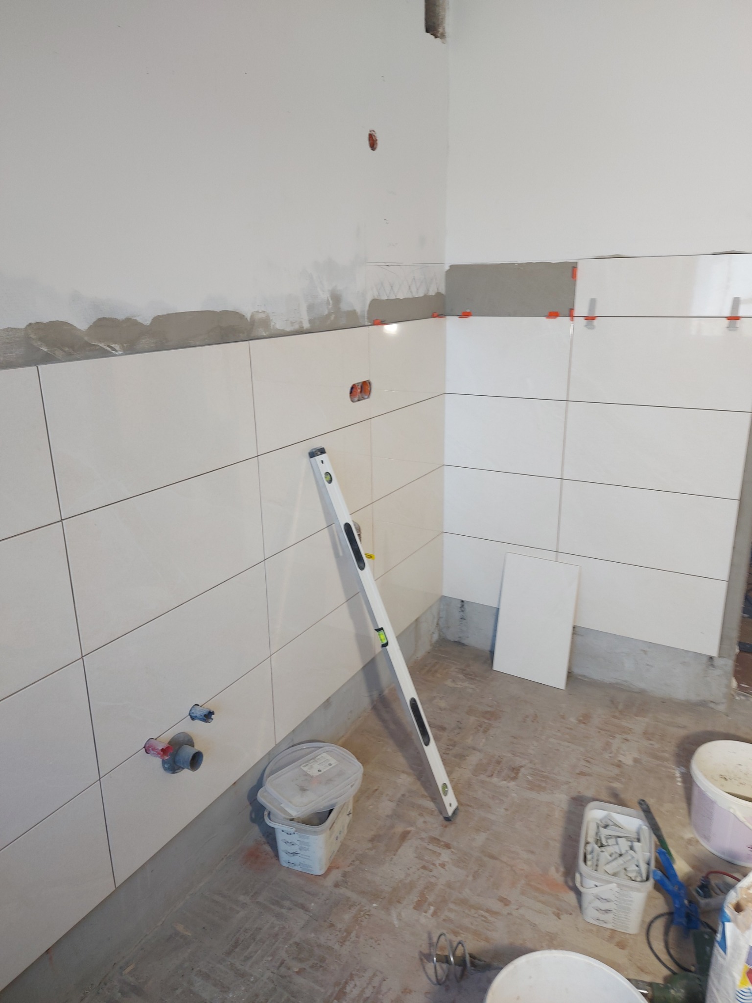 Białe, prostokątne płytki ceramiczne układane na ścianie, widoczne plastikowe krzyżyki dystansowe, poziomica oparta o ścianę, przygotowane rury instalacyjne, wiaderka i narzędzia na podłodze.
