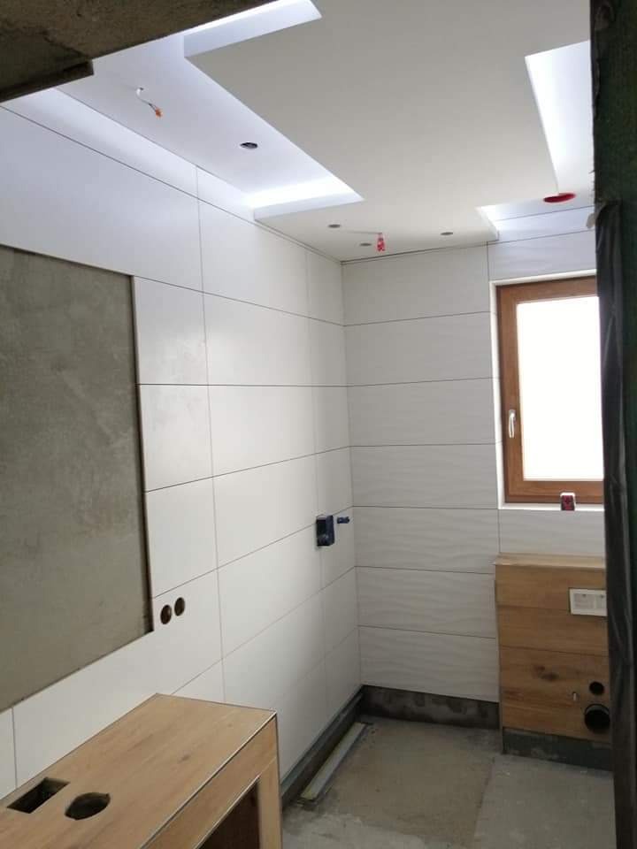 Wnętrze łazienki w trakcie prac wykończeniowych: białe, poziome płytki na ścianach, częściowo obudowane stelaże pod umywalkę i WC, podwieszany sufit z oświetleniem LED.