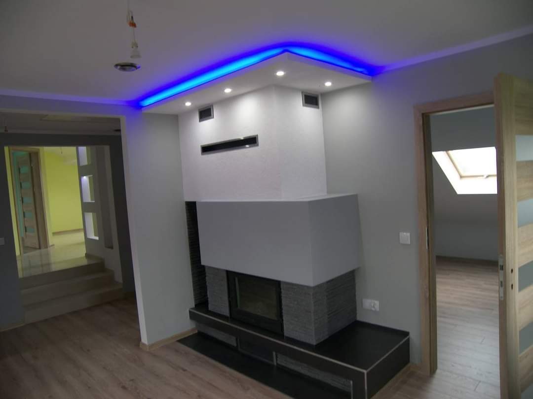 Nowoczesny salon z kominkiem obłożonym szarym kamieniem, podświetlany sufit z niebieskim LED, parkiet na podłodze i widok na poddasze przez otwarte drzwi.