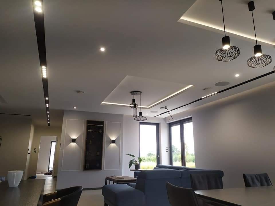 Nowoczesny salon z podwieszanym sufitem, oświetleniem punktowym i dekoracyjnymi lampami wiszącymi, szara sofa i ściany.