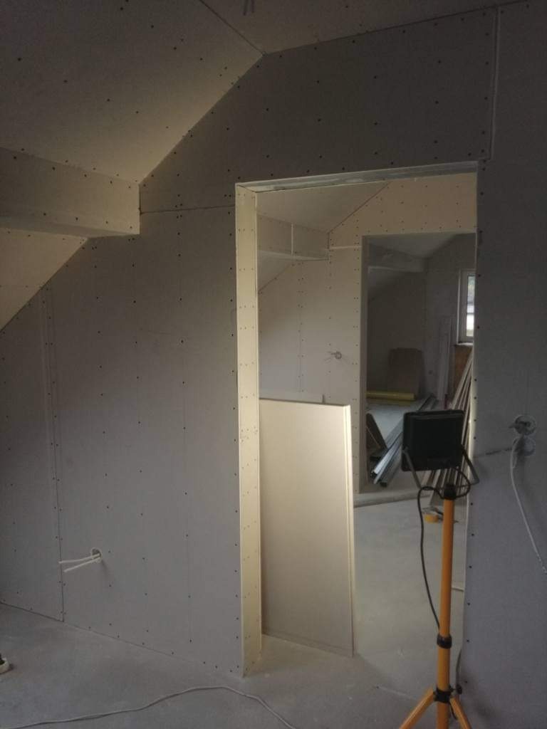 Wnętrze w trakcie prac wykończeniowych, ściany i sufit pokryte płytami gipsowo-kartonowymi, widoczna konstrukcja otworu drzwiowego i lampa budowlana na trójnogu.