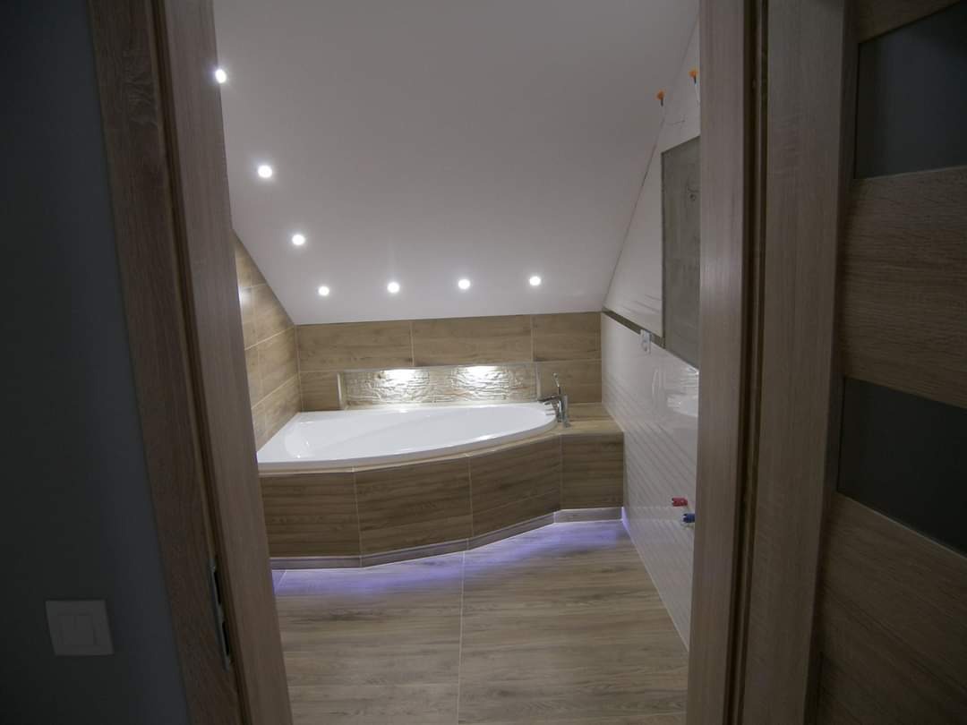 Wykończona łazienka na poddaszu z wanną w zabudowie z płytek imitujących drewno, oświetleniem punktowym i widocznym fragmentem drzwi z okleiną drewnopodobną.