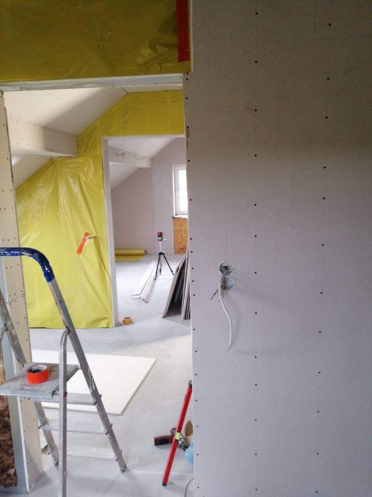 Montaż płyty gipsowo-kartonowej na poddaszu, widoczna drabina, poziomica i otwór na gniazdko elektryczne z wystającymi przewodami.