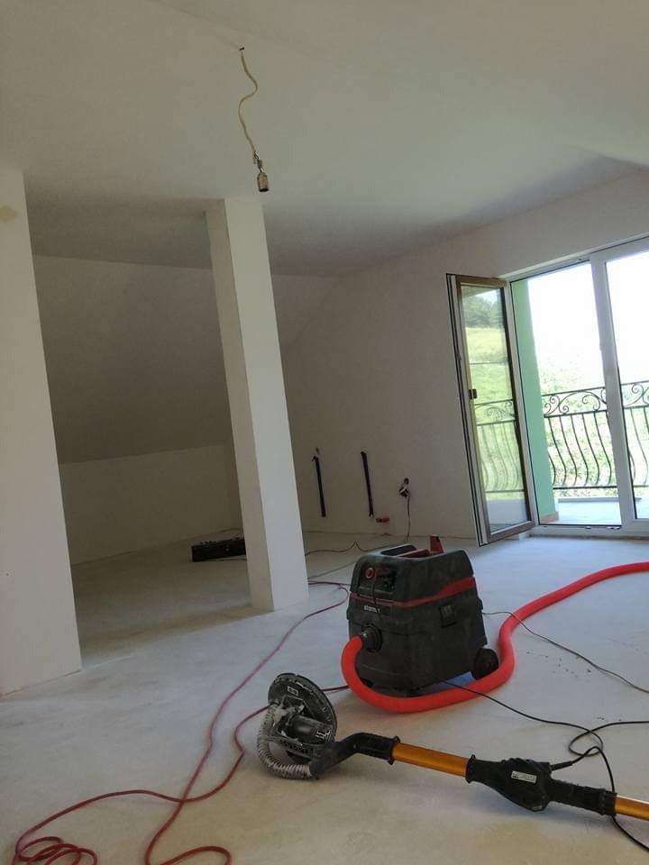 Wnętrze w trakcie prac wykończeniowych: szlifierka do gładzi, odkurzacz przemysłowy i surowa żarówka zwisająca z sufitu w pokoju z oknem balkonowym.