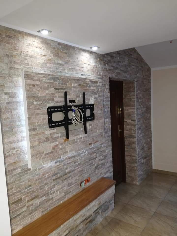 Ściana wykończona kamieniem dekoracyjnym w odcieniach beżu i szarości, z zamontowanym uchwytem do telewizora i drewnianą półką, oświetlona punktowymi lampami sufitowymi.