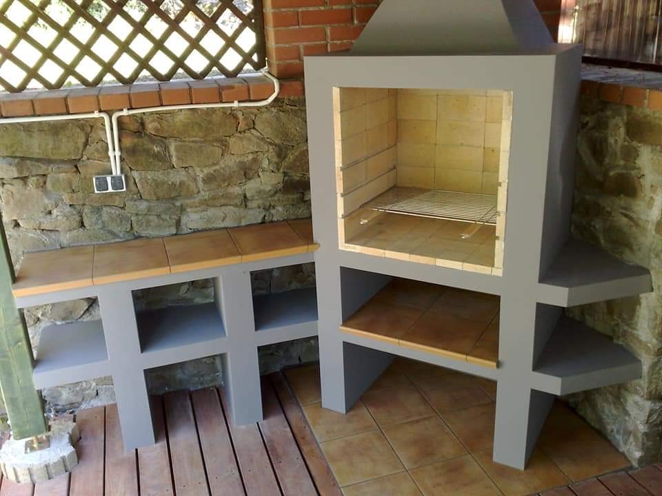 Szary murowany grill z blatem wyłożonym płytkami, z półkami i rusztem, na zewnątrz, na drewnianym tarasie, obok ściany z kamienia.