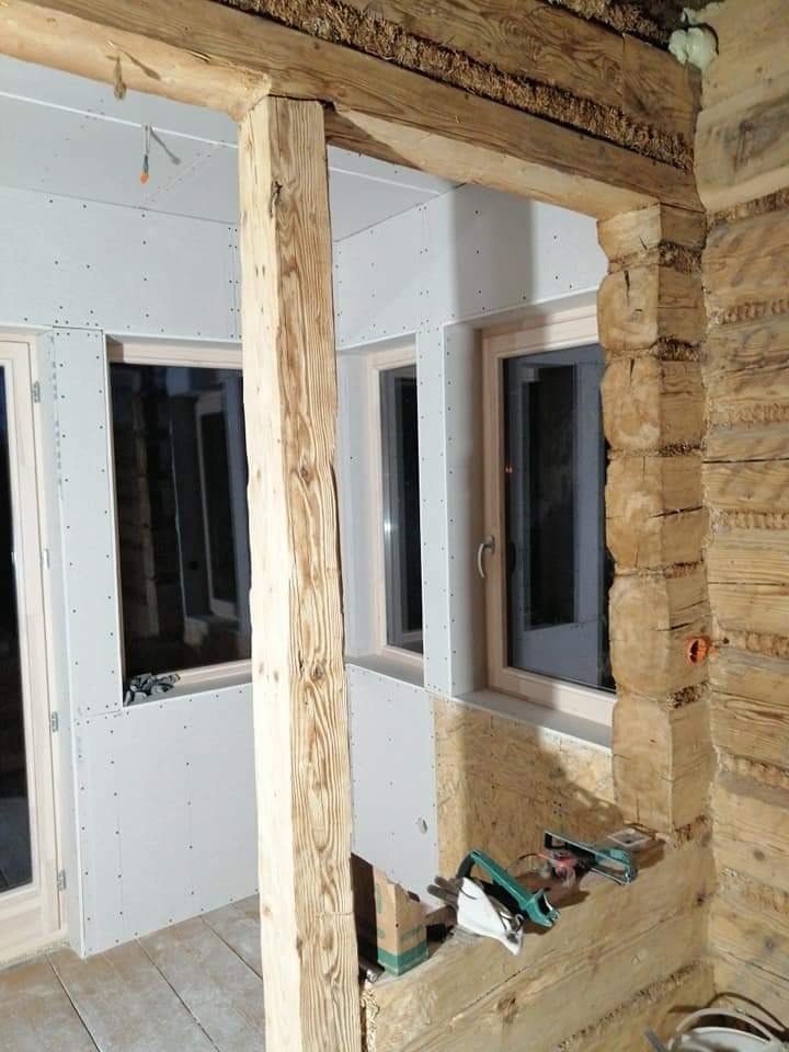 Montaż płyt karton gips na ścianach w drewnianym domu z widocznymi belkami stropowymi, narzędzia do cięcia płyt leżą na podłodze.