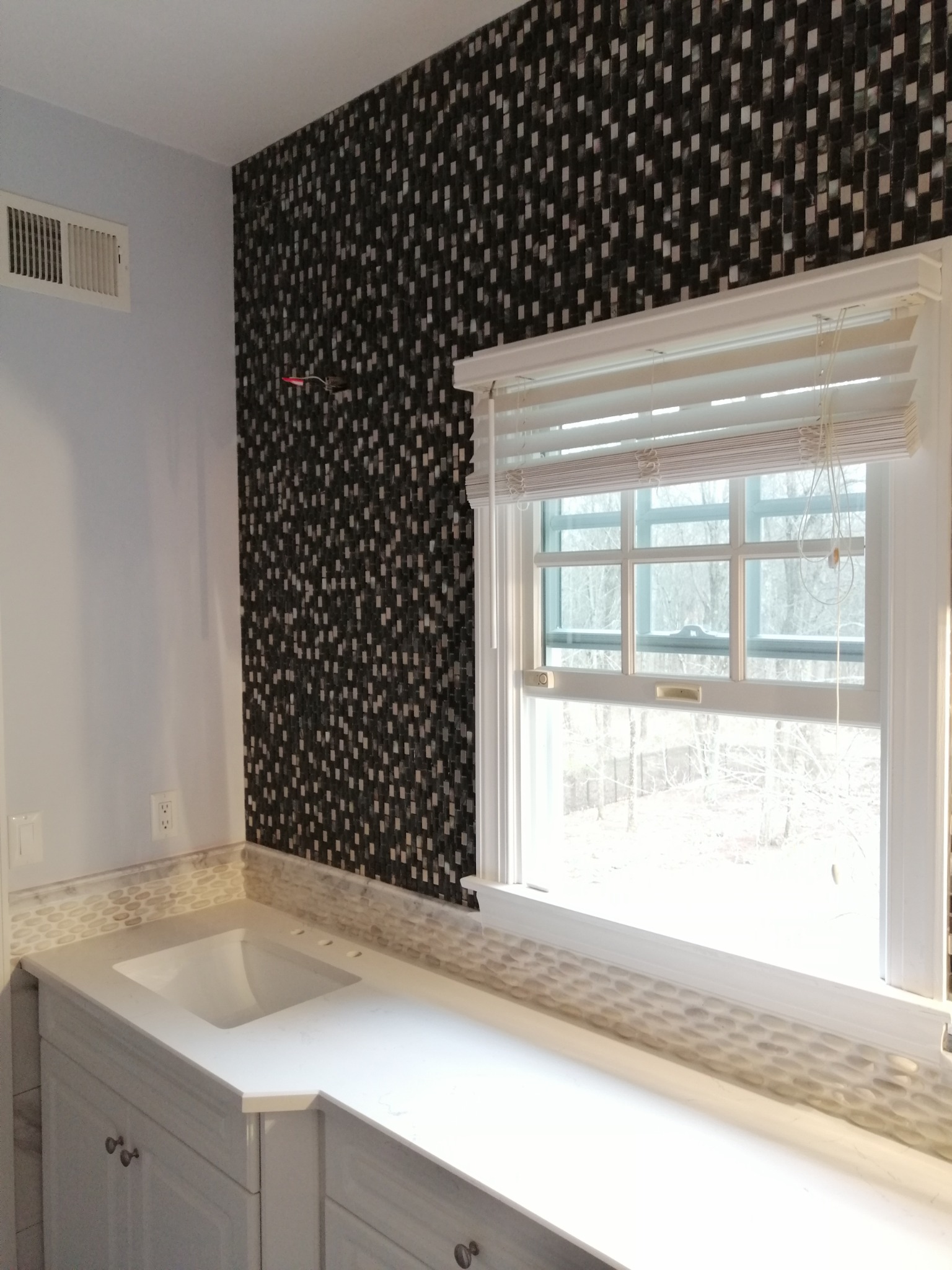 Nowoczesna łazienka z czarno-białą mozaiką na ścianie, białą szafką pod umywalką i blatem z jasnego konglomeratu oraz oknem z białymi żaluzjami.