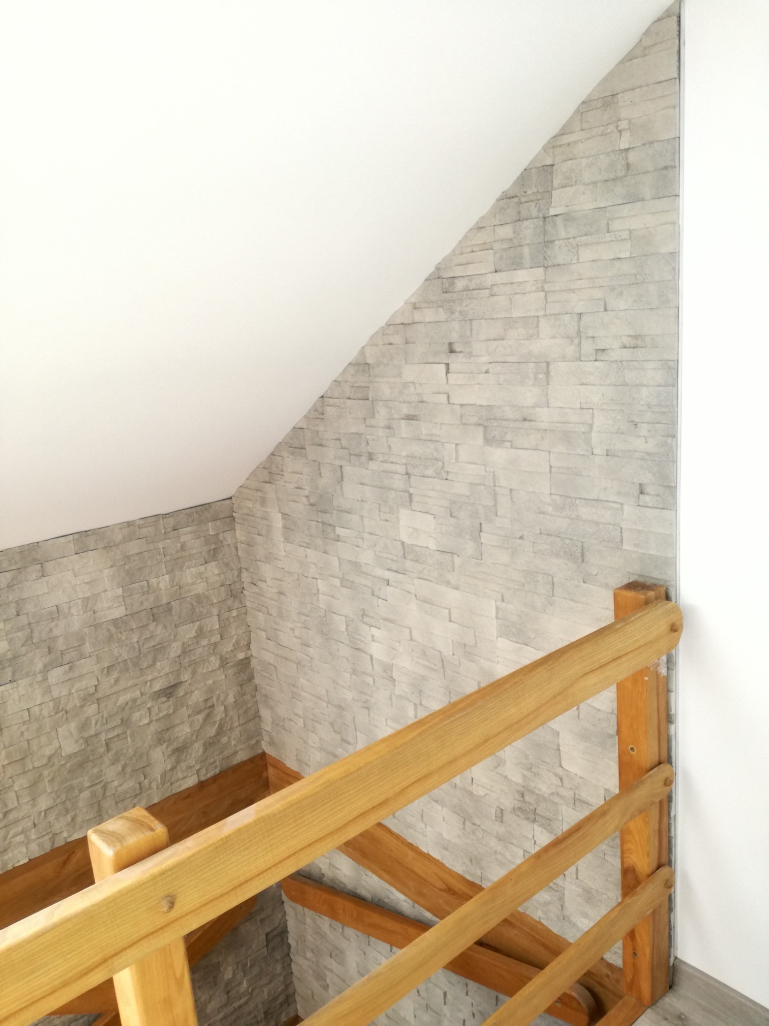 Ściana wykończona szarą okładziną imitującą kamień w poddaszowym pomieszczeniu z drewnianą barierką w Myślenicach.