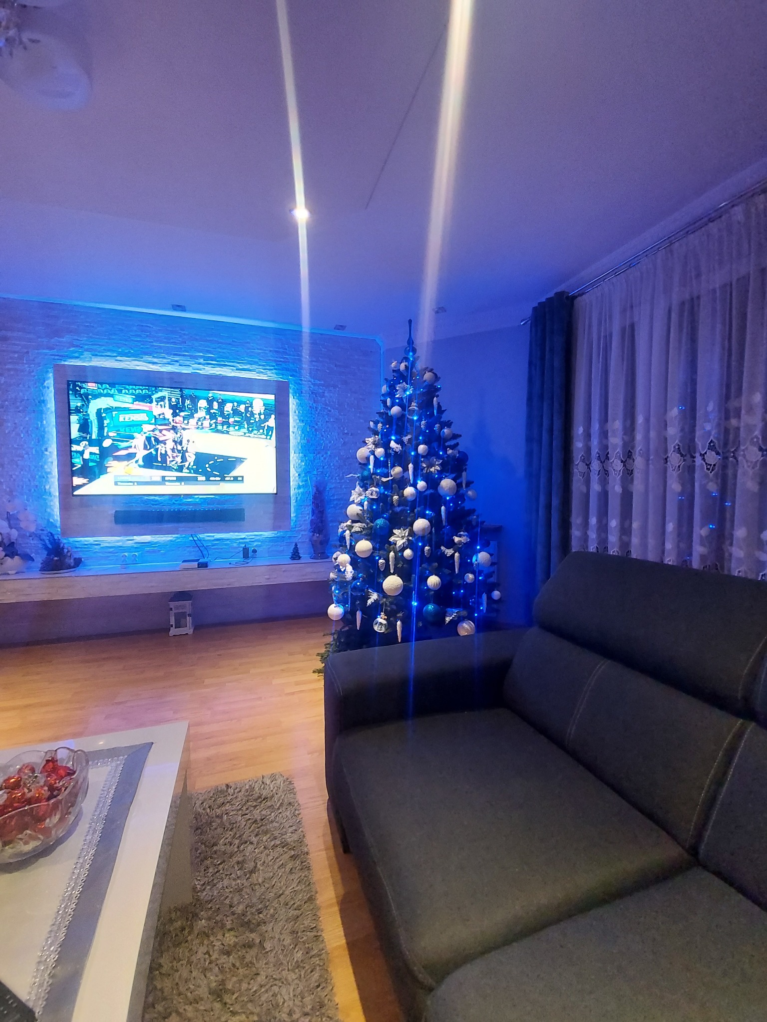 Salon z podświetlanym telewizorem na ścianie z imitacji kamienia, choinką z białymi bombkami i ciemną sofą, oświetlony niebieskim światłem.