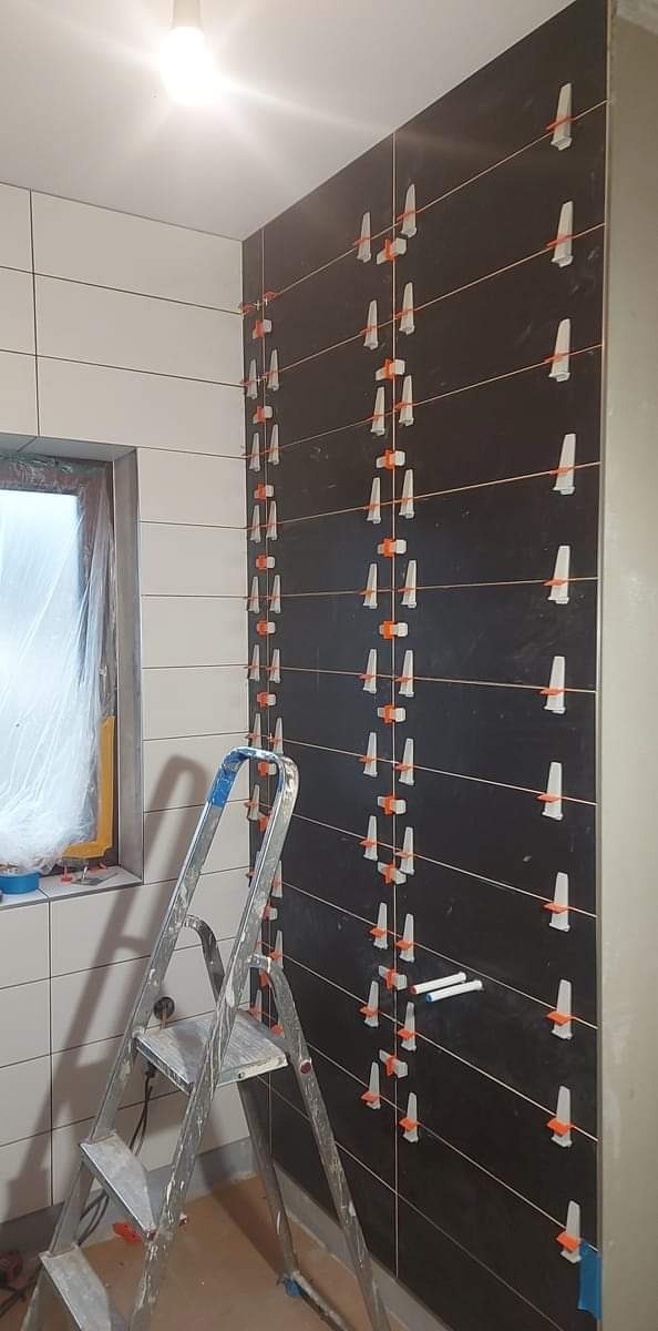 Układanie ciemnych płytek na ścianie w łazience, widoczne poziomice i klipsy do płytek, drabina aluminiowa w tle, okno z folią ochronną.