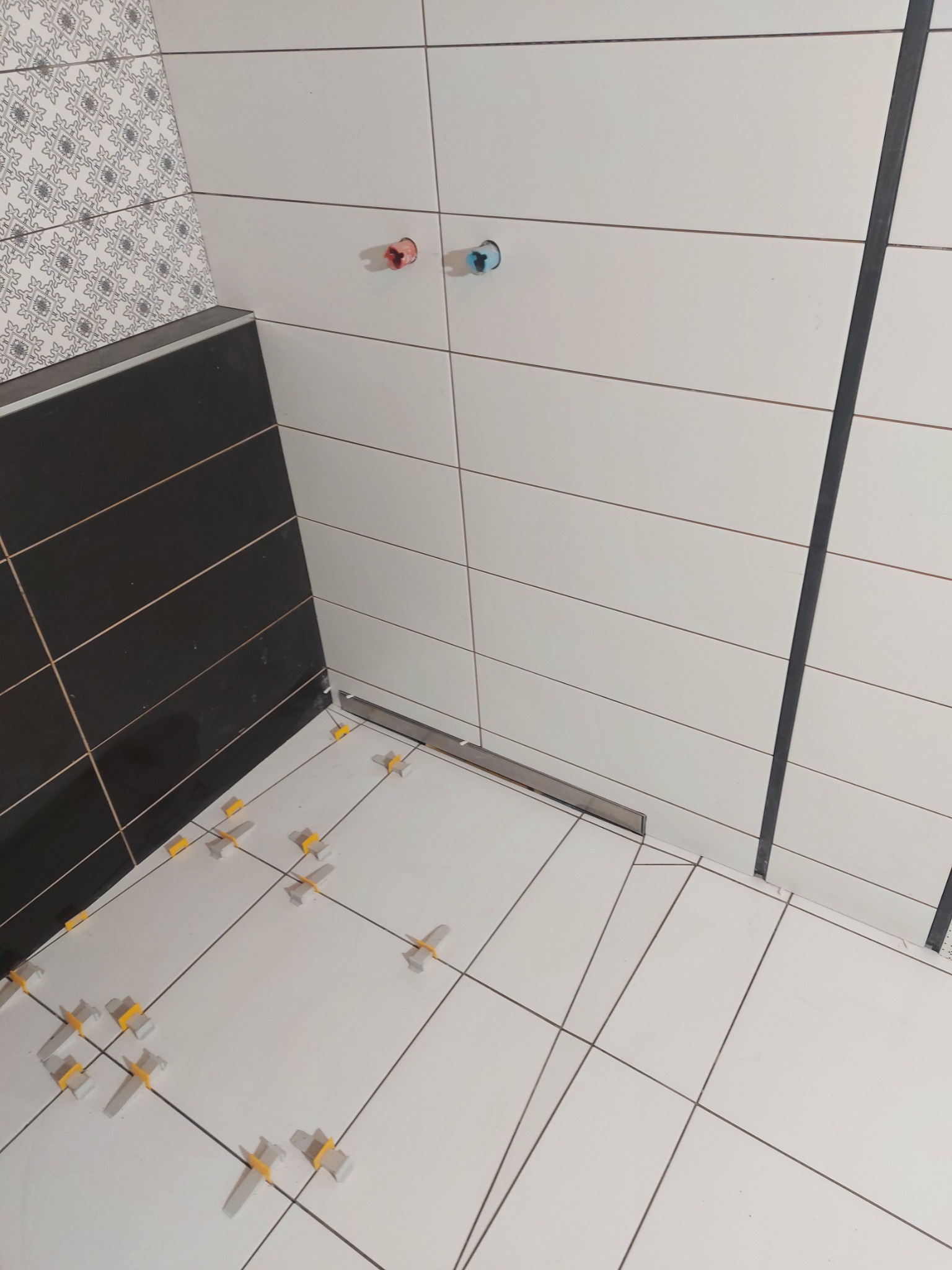 Biało-czarna łazienka w trakcie wykańczania: białe płytki podłogowe i ścienne z krzyżykami dystansowymi, czarne płytki na ścianie, widoczne odpływy liniowe.