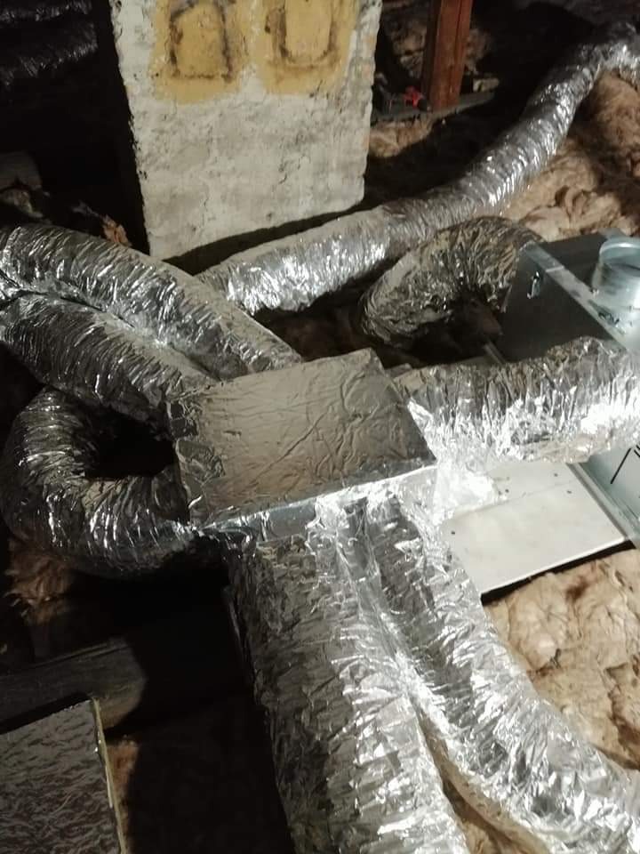 Srebrne, elastyczne rury wentylacyjne połączone kwadratowym elementem, ułożone na izolacji z wełny mineralnej, widoczne fragmenty starego komina z żółtymi śladami po naprawach.