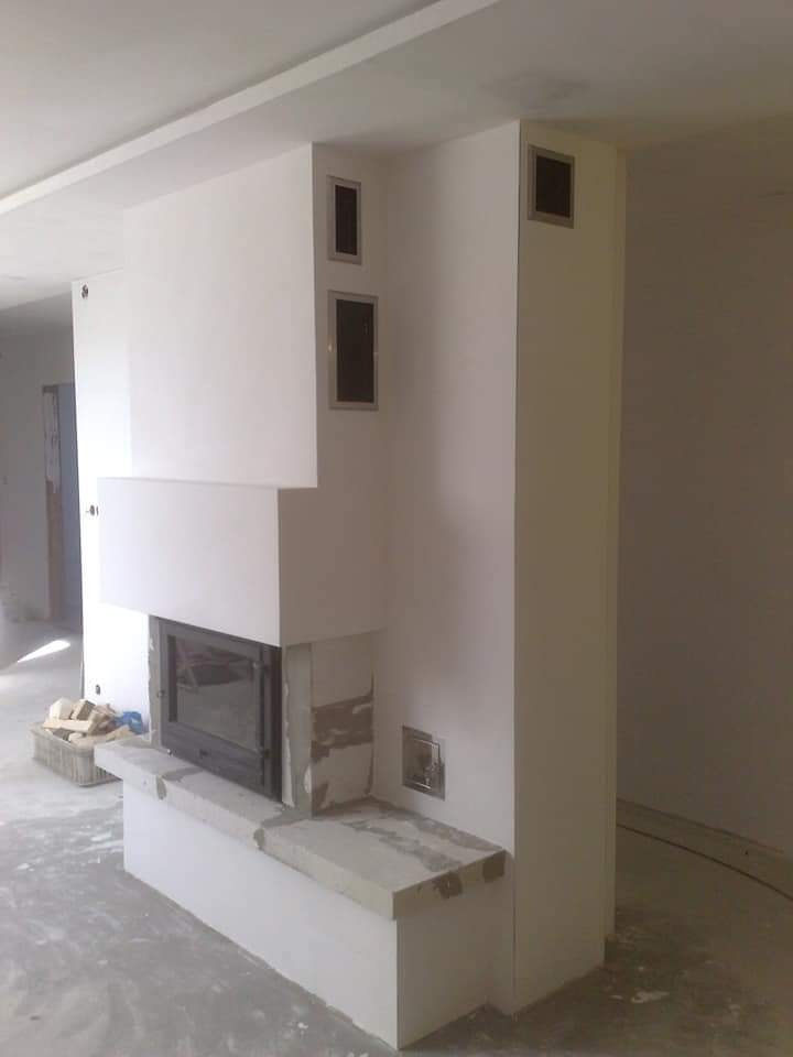 Wnętrze w trakcie prac: zabudowa kominka z wkładem, surowy beton komórkowy i białe płyty gipsowo-kartonowe, widoczne kratki wentylacyjne w obudowie.