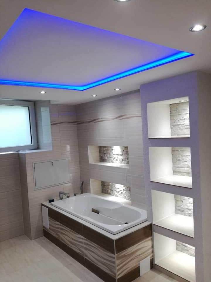 Nowoczesna łazienka z podświetlanym sufitem LED, beżowymi płytkami i wnękami dekoracyjnymi wyłożonymi kamieniem, wanna obudowana płytkami z motywem fal.