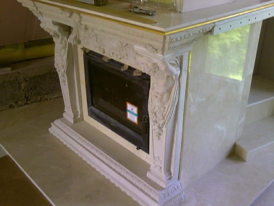 Wykończony obudowany kominek z dekoracyjnymi rzeźbieniami i marmurowym wykończeniem, z widocznym fragmentem podłogi i ściany w tle.