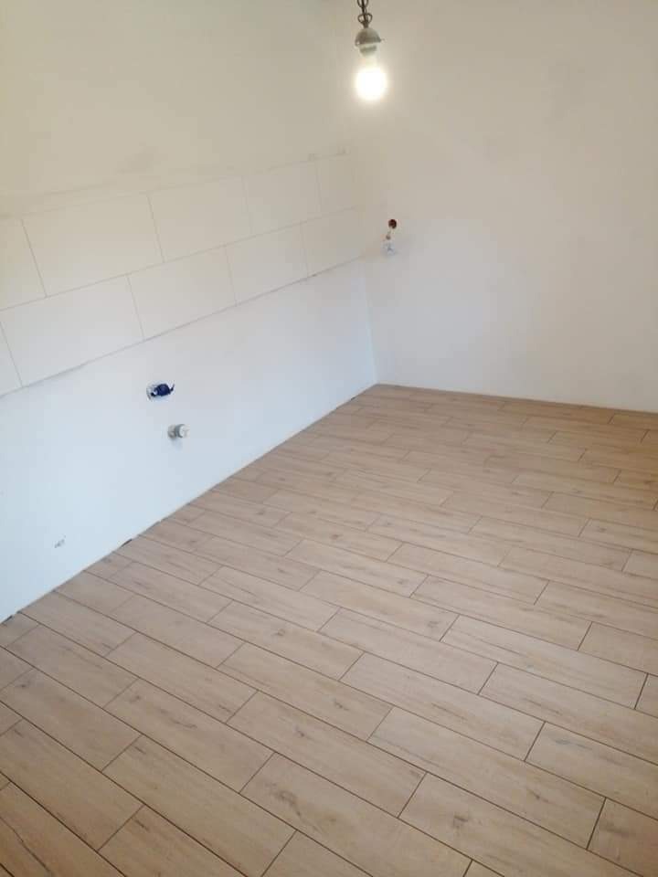 Wnętrze po częściowym remoncie: ułożona podłoga z paneli imitujących drewno, na ścianie częściowo położone białe płytki. Widoczne wystające kable i instalacje.