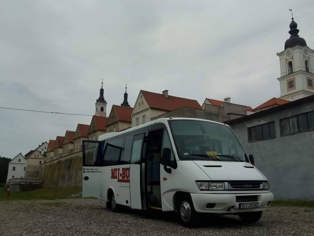 Biały bus marki Iveco z otwartymi drzwiami na tle zabudowań klasztornych z wieżą zegarową.