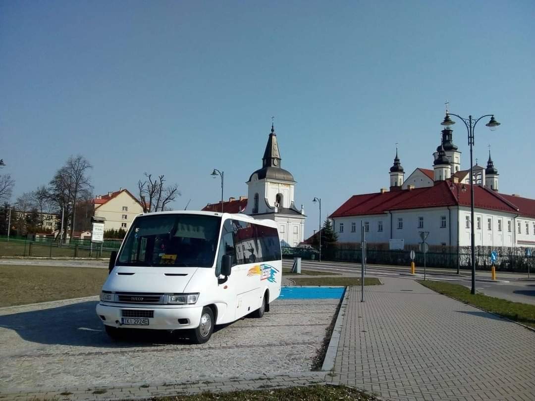 Biały bus Iveco na parkingu z kostki brukowej, w tle budynki sakralne z czerwonymi dachami i wieżami, błękitne niebo.