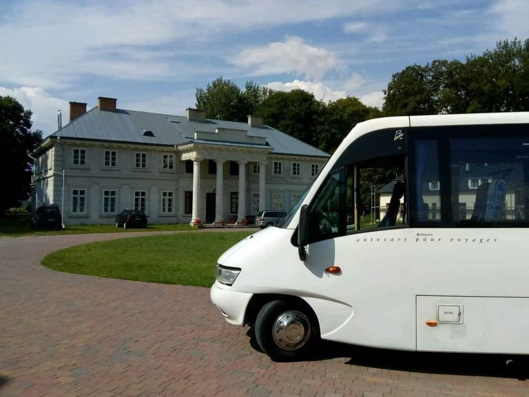 Biały bus turystyczny na brukowanym podjeździe przed klasycystycznym budynkiem z kolumnami.