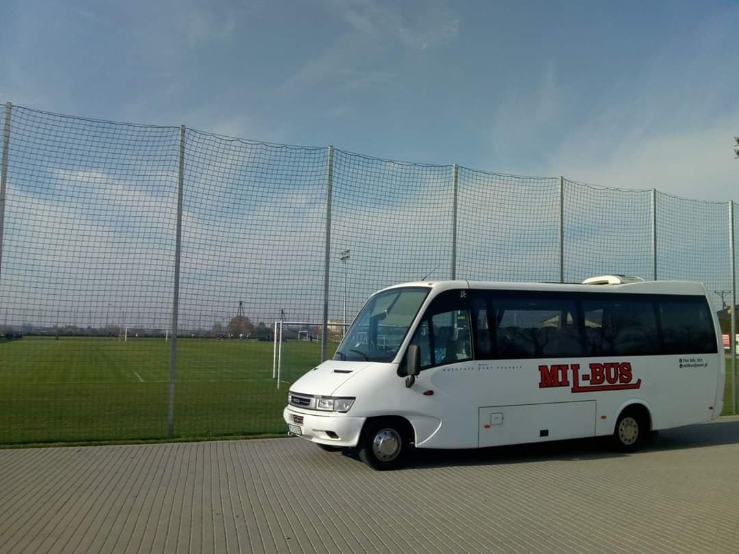 Biały minibus z czerwonym napisem 'MIL-BUS' zaparkowany na chodniku, w tle siatka ogrodzeniowa i boisko sportowe.