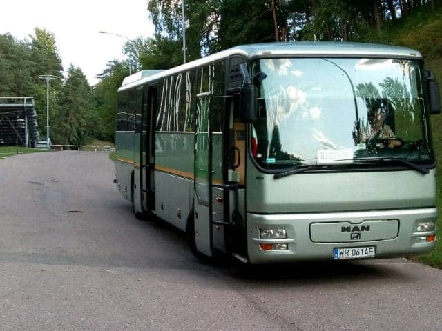 Srebrny autobus MAN z otwartymi drzwiami, zaparkowany na asfaltowej drodze na tle zieleni, widoczny kierowca w odbiciu szyby.