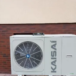 Zewnętrzna jednostka pompy ciepła Kaisai ustawiona na metalowej konstrukcji przy ścianie budynku z poziomnicą na górze, sprawdzana podczas instalacji.