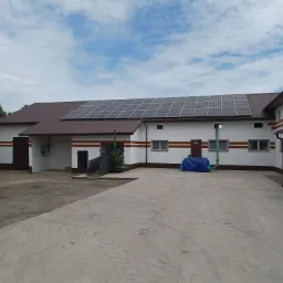 Biały budynek z brązowym dachem i panelami słonecznymi, widok z zewnątrz na betonowy plac w słoneczny dzień.