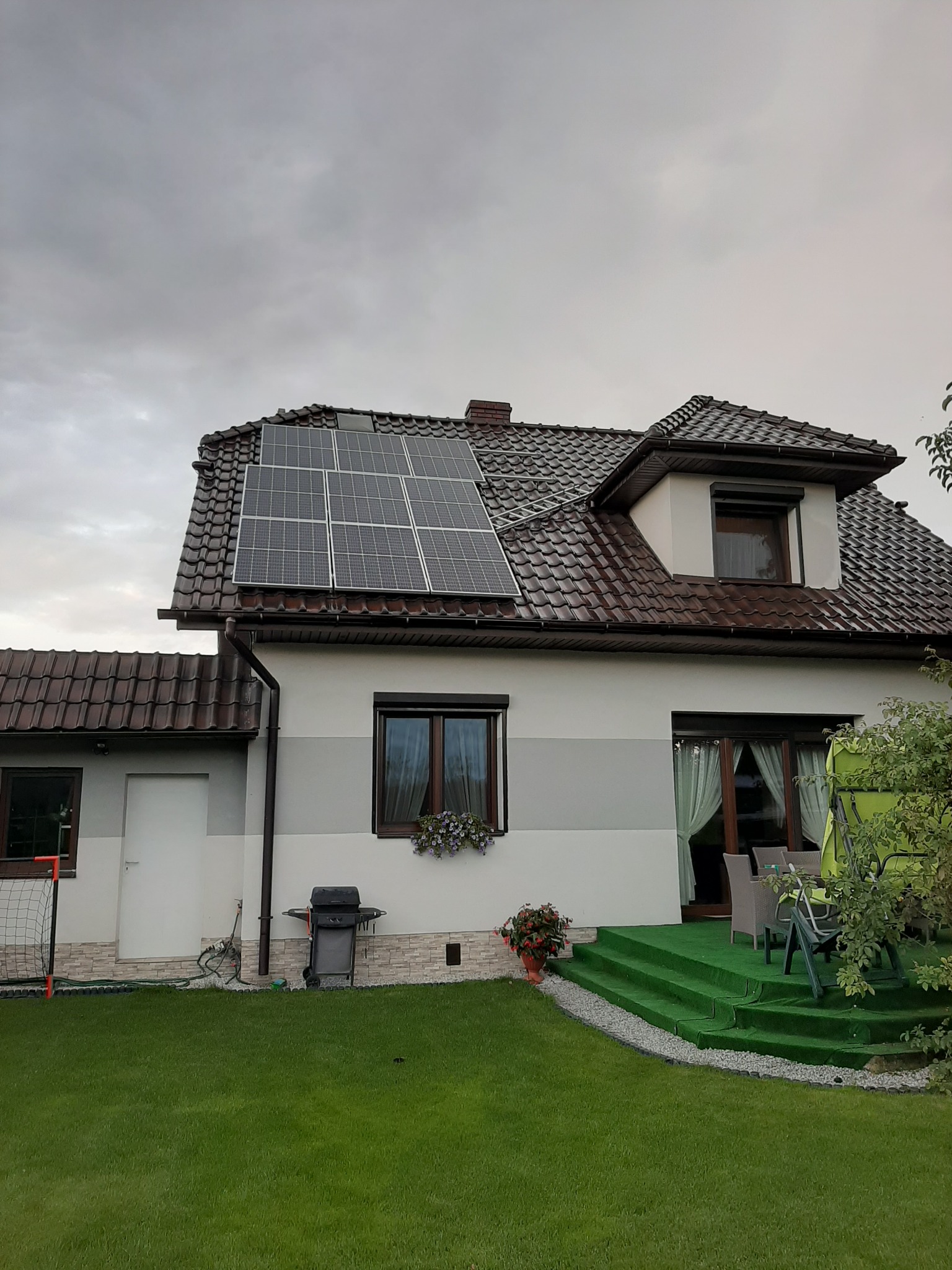 Dom jednorodzinny z brązową dachówką i panelami fotowoltaicznymi na dachu, widok z trawnika w pochmurny dzień.