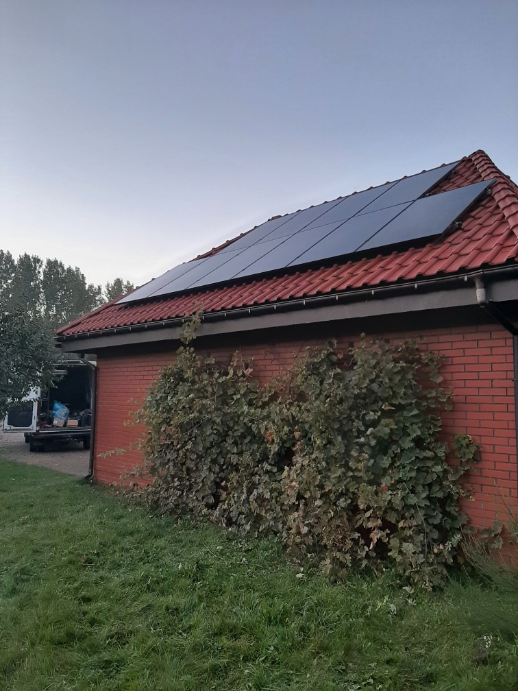 Panele fotowoltaiczne na dachu domu z czerwonej cegły, częściowo zakryte przez pnącą roślinność, z widocznym samochodem dostawczym w tle.