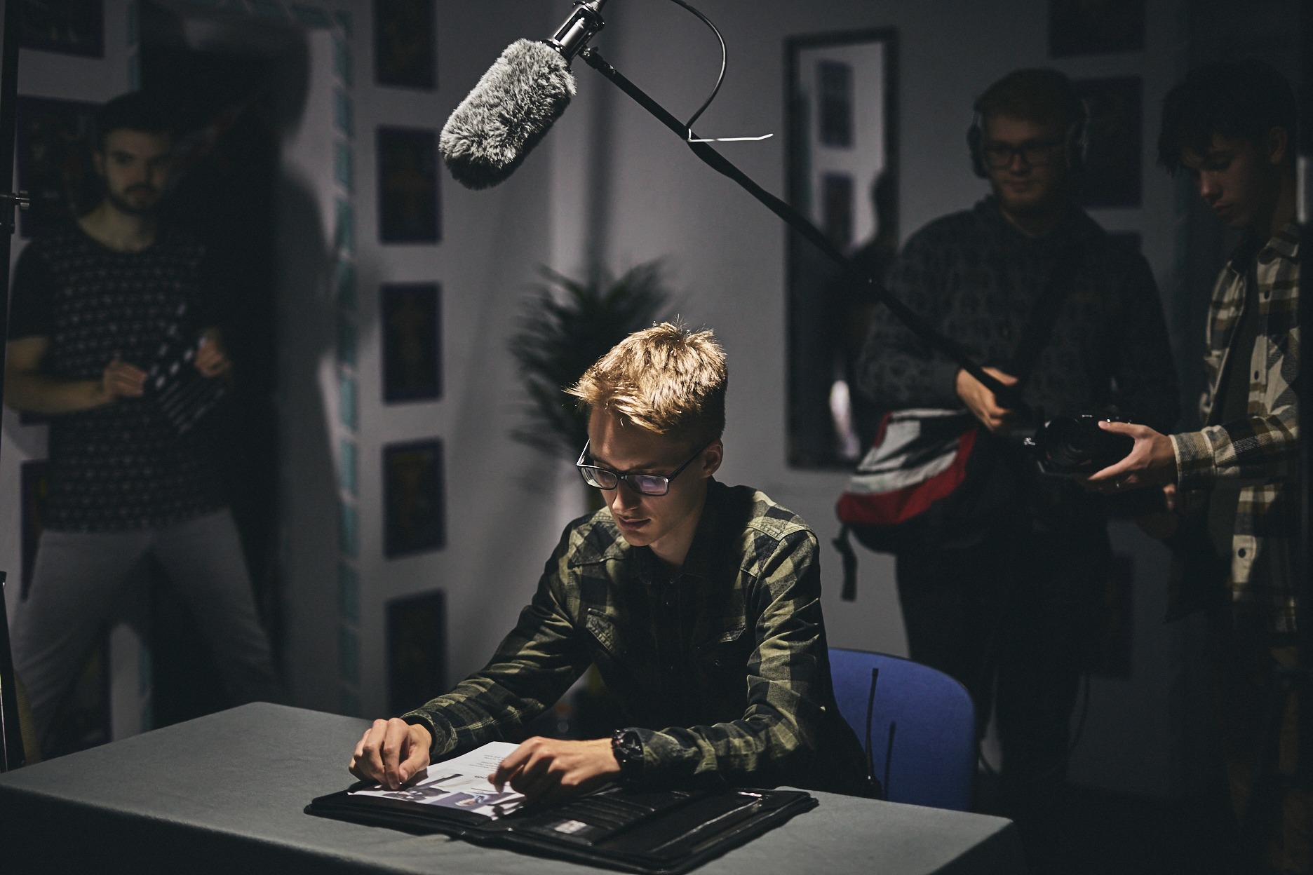Młody mężczyzna w okularach przegląda portfolio przy stole w studiu filmowym, z mikrofonem na wysięgniku i ekipą filmową w tle.