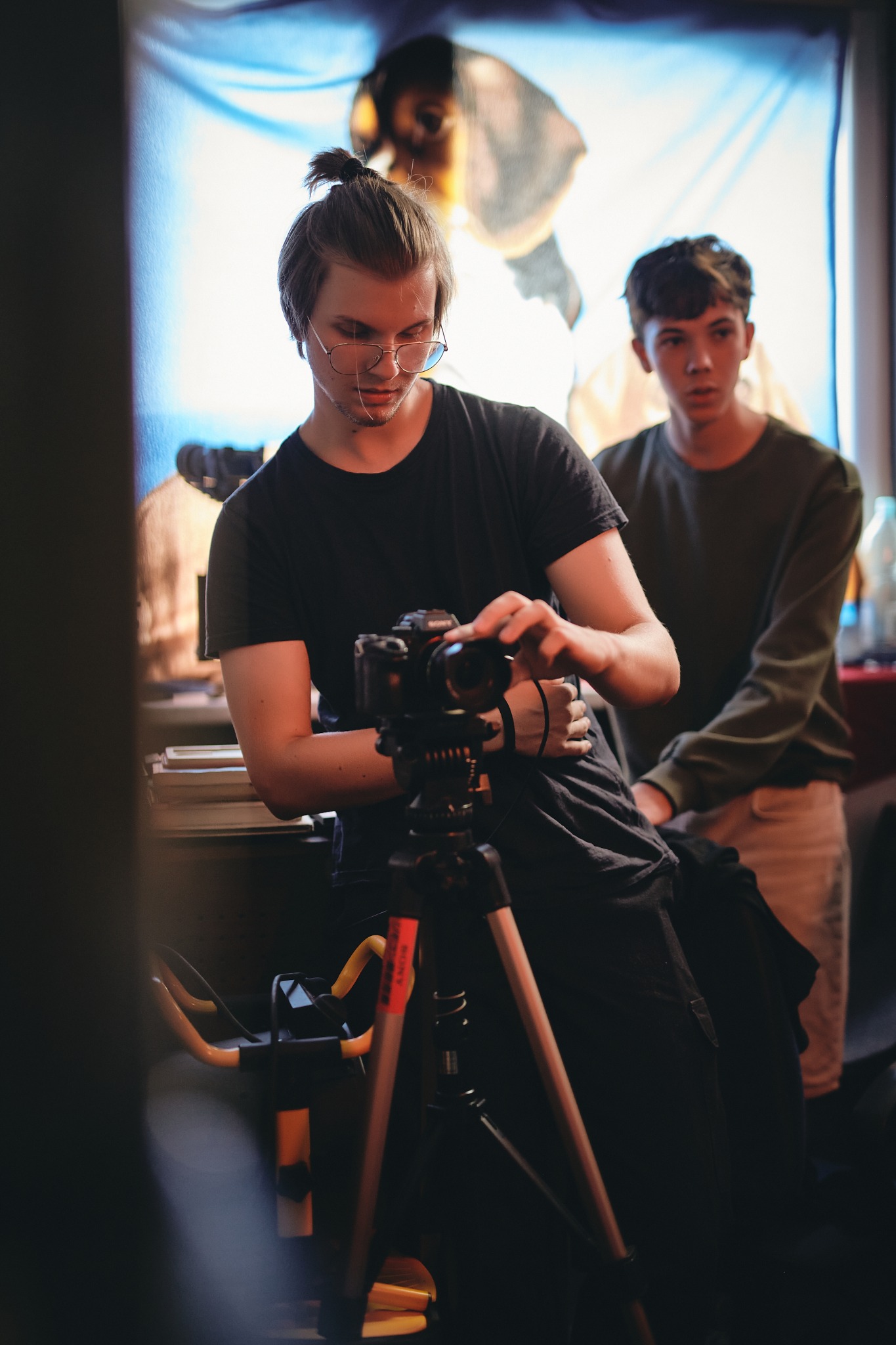 Operator kamery w okularach, ustawia ostrość, drugi mężczyzna obserwuje, w tle rozmyta grafika, Warszawa.