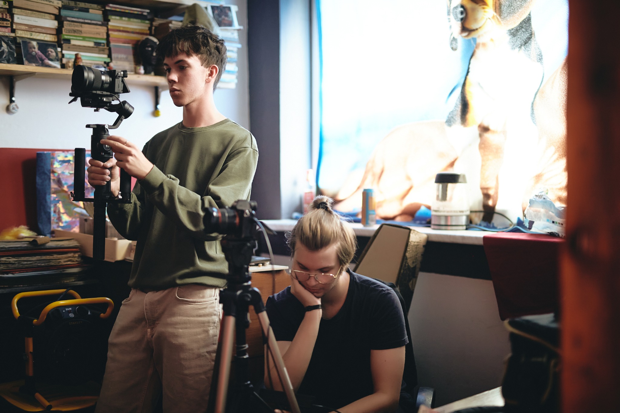 Dwóch młodych mężczyzn w pomieszczeniu z rekwizytami filmowymi: jeden obsługuje kamerę na stabilizatorze, drugi siedzi przy statywie, obok okna z pluszowym psem.