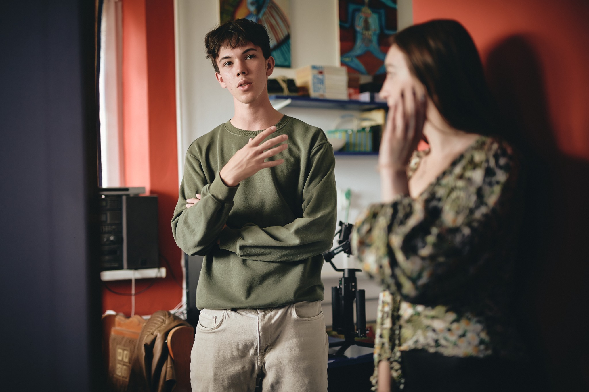 Młody mężczyzna w oliwkowej bluzie gestykuluje podczas rozmowy z kobietą w kwiecistej bluzce, w tle widoczny sprzęt do filmowania.