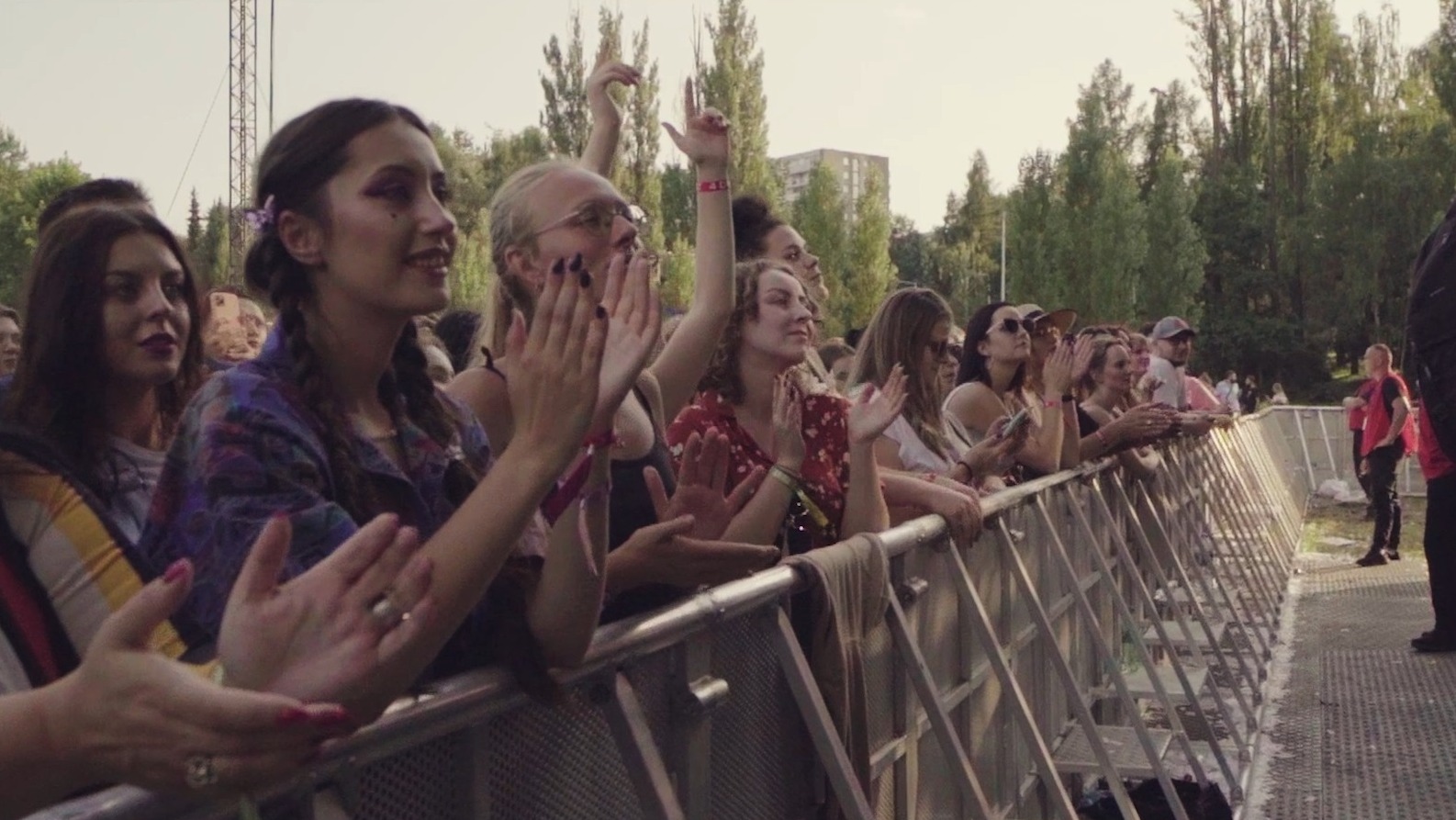 Widok zza barierek na publiczność klaszczącą podczas koncertu plenerowego, z widocznymi drzewami i budynkiem w tle.