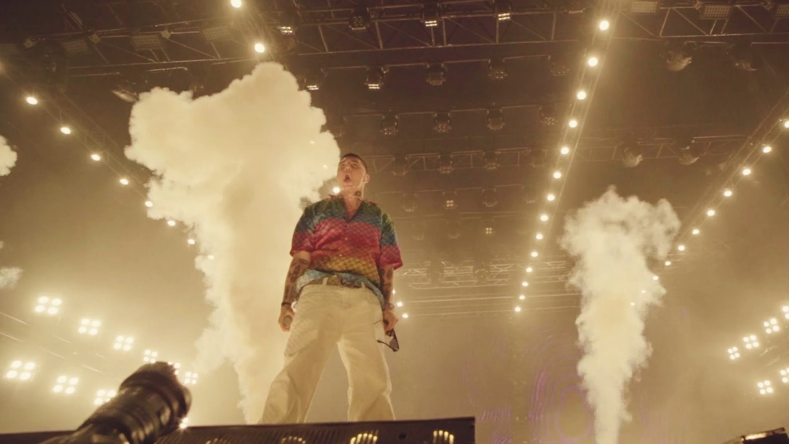 Piosenkarz w koszuli w paski na scenie z wytwornicami dymu i oświetleniem koncertowym.