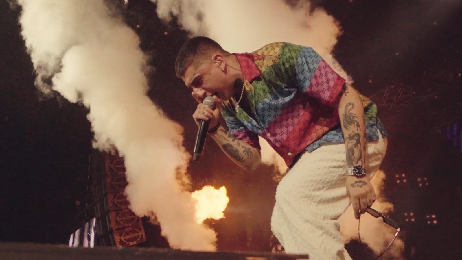 Wokalista z tatuażami na ramionach, w kolorowej koszuli i białych spodniach, śpiewa do mikrofonu na scenie z efektami pirotechnicznymi i dymem.