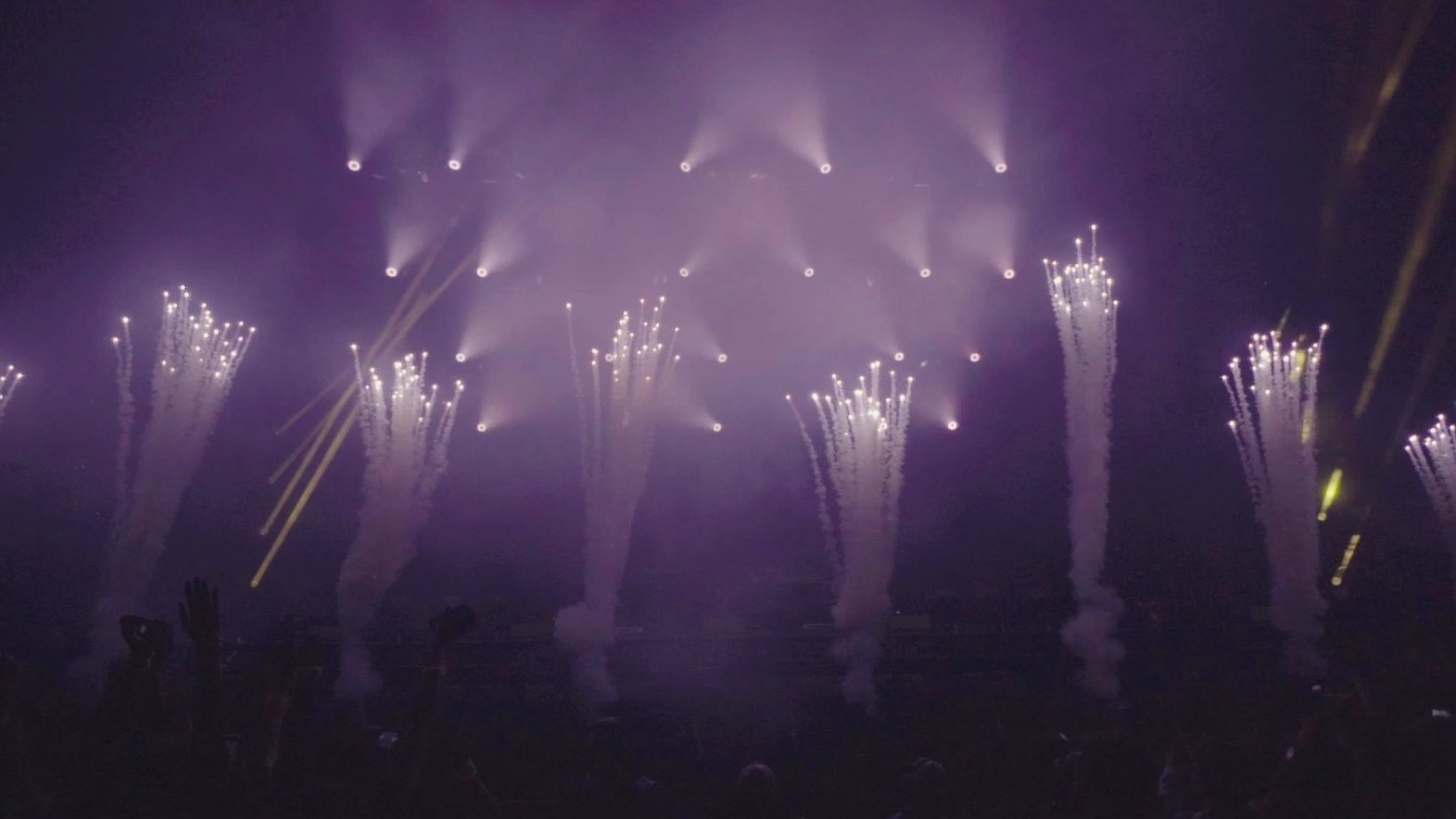 Scena koncertowa w fioletowym oświetleniu z pionowymi strumieniami iskier i rozmazanymi sylwetkami publiczności w dolnej części kadru.