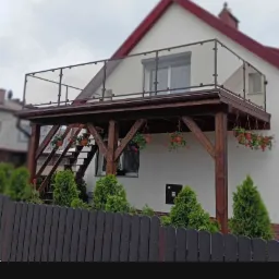 Zadaszony balkon z drewnianą konstrukcją wsporczą, szklaną balustradą i schodami, ozdobiony wiszącymi donicami z czerwonymi kwiatami, widoczny zza ciemnego drewnianego ogrodzenia i zielonych krzewów.