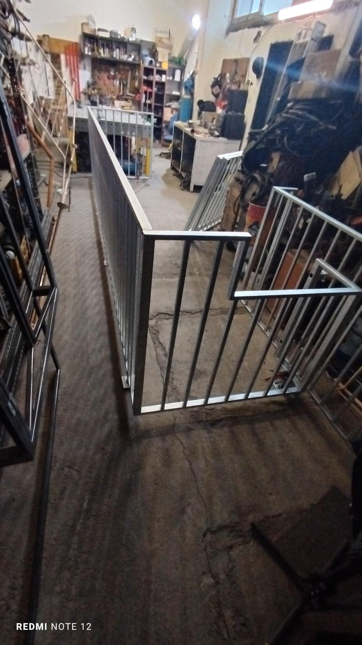Wnętrze warsztatu z metalową balustradą schodową w trakcie produkcji, widoczne surowe wykończenie i narzędzia w tle.