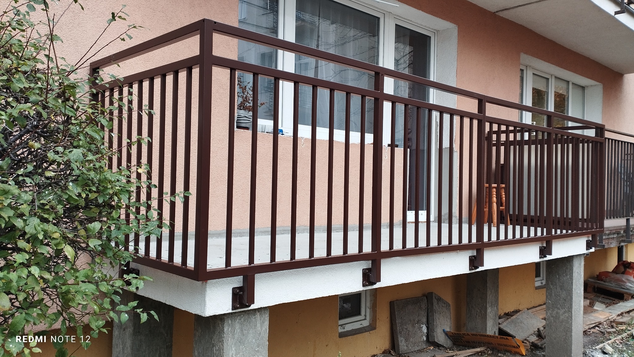 Balkon z brązową metalową balustradą z pionowymi prętami, widok z zewnątrz, na pierwszym planie krzew z zielonymi liśćmi.