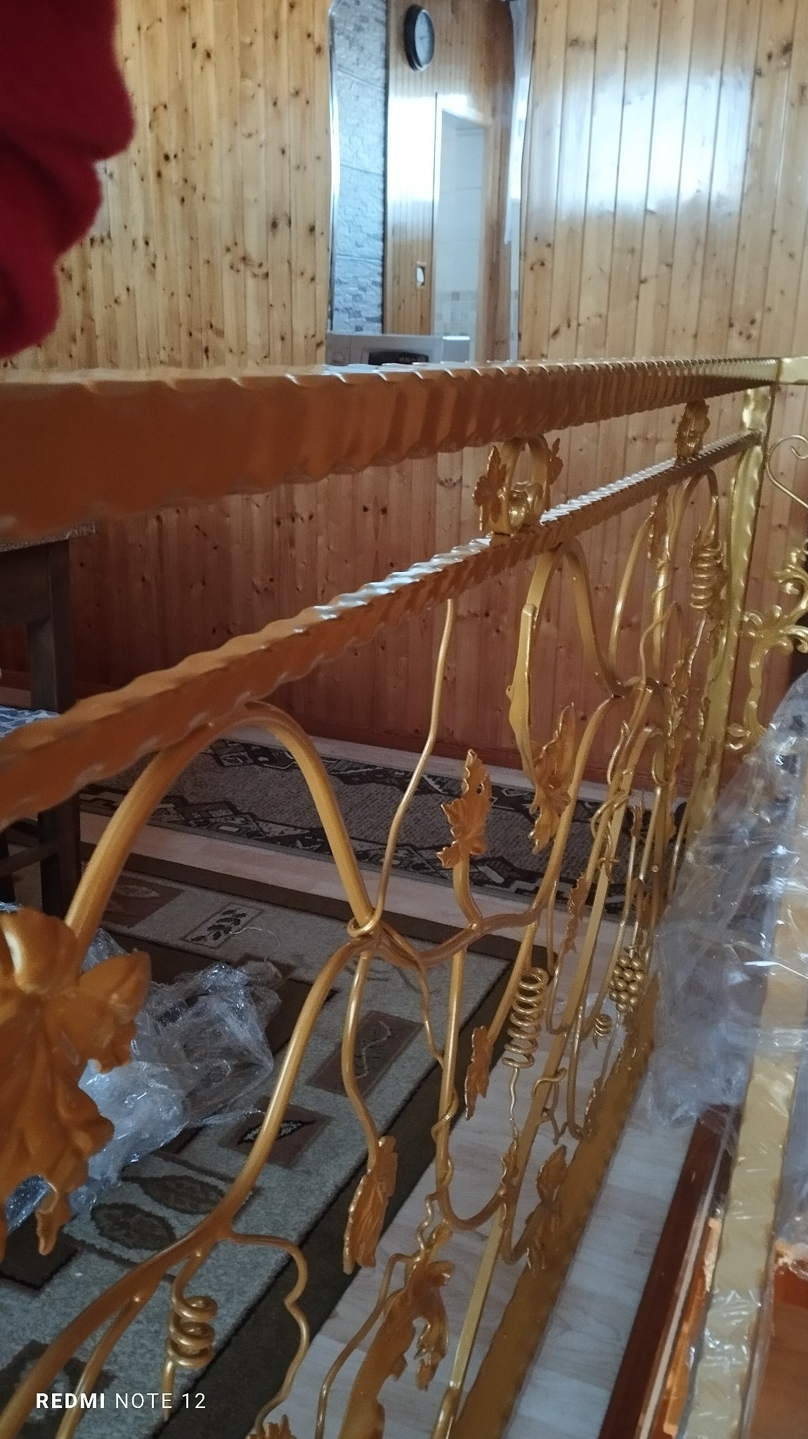 Złota balustrada z motywem roślinnym, widoczna w rustykalnym wnętrzu z boazerią i dywanem w geometryczne wzory. Elementy balustrady owinięte folią ochronną.