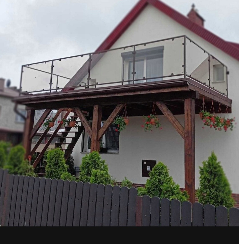 Zadaszony balkon z drewnianą konstrukcją wsporczą, szklaną balustradą i schodami, ozdobiony wiszącymi donicami z czerwonymi kwiatami, widoczny zza ciemnego drewnianego ogrodzenia i zielonych krzewów.