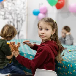 Dziewczynka w czerwonej bluzie trzyma pomalowaną, glinianą figurkę zwierzęcia na tle stołu z kolorowym obrusem i balonami, podczas zajęć plastycznych.