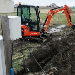 Pomarańczowa mini koparka Kubota KX019-4 na grząskim terenie wykopuje wąski r&oacute;w, obok skrzynka elektryczna i kable, w tle domy jednorodzinne.