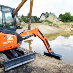 Pomarańczowa mini koparka Kubota KX027-4 z czarną łyżką zanurzoną w wodzie podczas kopania stawu, w tle dom i hałda piasku.