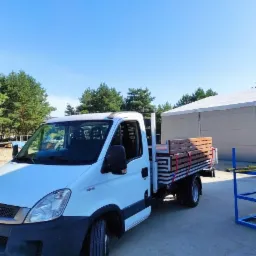 Biały samochód dostawczy Iveco z platformą załadowaną drewnianymi elementami, zabezpieczonymi czerwonymi pasami, na tle błękitnego nieba i zielonych drzew.