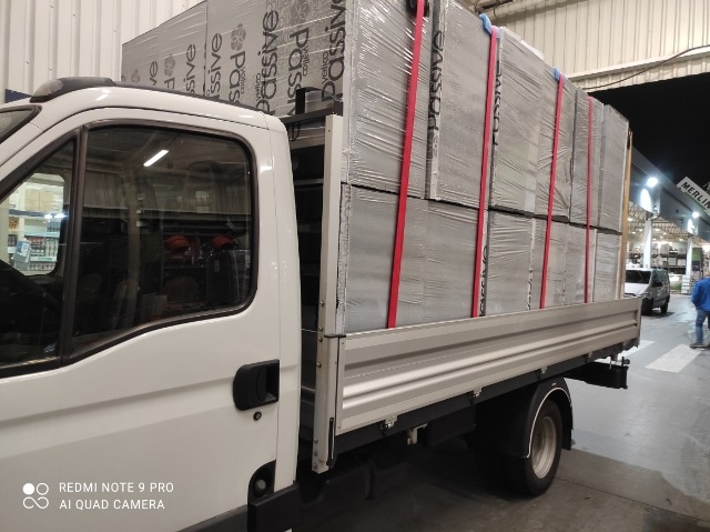 Biały samochód dostawczy z platformą załadowaną płytami izolacyjnymi zabezpieczonymi czerwonymi pasami transportowymi, widok z boku na tle budynku magazynowego.