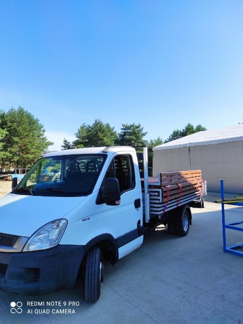 Biały samochód dostawczy Iveco z platformą załadowaną drewnianymi elementami, zabezpieczonymi czerwonymi pasami, na tle błękitnego nieba i zielonych drzew.